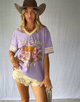 A woman modeling a lavender purple graphic t-shirt with yellow and white trim, paired with a brown cowboy hat, beige shorts, and green boots against a light gray backdrop.
