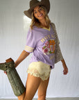 A woman modeling a lavender v-neck graphic t-shirt with baby yellow stripes on the sleeves and a matching baby yellow v neck collar, paired with a brown cowboy hat, yellow lace trim shorts and green cowboy boots. The graphic tee reads "Hot Girl Juice" and is a playful nod to spicy marg lovers. The perfect dopamine dressing tee for the modern coastal cowgirl.