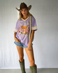 A woman modeling a lavender purple graphic t-shirt with a western theme, paired with denim shorts, a brown cowboy hat, and green cowboy boots. Modern coastal cowgirl.