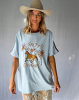 A woman wearing a light blue graphic t-shirt with a cowboy hat and a white lace and sequin skirt. The oversized tee features a hand-drawn graphic of a cowgirl in a shell bikini top riding a horse, palm trees, and the text 'SALTY COWGIRL'. For the modern salty cowgirl, this iconic tee is perfect for festival styling, rodeos and everyday wear all year round.