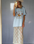 A woman wearing a light blue graphic t-shirt with a cowboy hat and a white lace and sequin skirt. The oversized tee features a hand-drawn graphic of a cowgirl in a shell bikini top riding a horse, palm trees, and the text 'SALTY COWGIRL'. For the modern salty cowgirl, this iconic tee is perfect for festival styling, rodeos and everyday wear all year round.