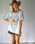 A woman wearing a light blue graphic t-shirt with a cowboy hat and a white lace and sequin skirt. The oversized tee features a hand-drawn graphic of a cowgirl in a shell bikini top riding a horse, palm trees, and the text 'SALTY COWGIRL'. For the modern salty cowgirl, this iconic tee is perfect for festival styling, rodeos and everyday wear all year round.
