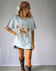 A woman wearing a light blue graphic t-shirt with a cowboy hat and a white lace and sequin skirt. The oversized tee features a hand-drawn graphic of a cowgirl in a shell bikini top riding a horse, palm trees, and the text 'SALTY COWGIRL'. For the modern salty cowgirl, this iconic tee is perfect for festival styling, rodeos and everyday wear all year round.
