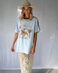 A woman wearing a light blue graphic t-shirt with a cowboy hat and a white lace and sequin skirt. The oversized tee features a hand-drawn graphic of a cowgirl in a shell bikini top riding a horse, palm trees, and the text 'SALTY COWGIRL'. For the modern salty cowgirl, this iconic tee is perfect for festival styling, rodeos and everyday wear all year round.