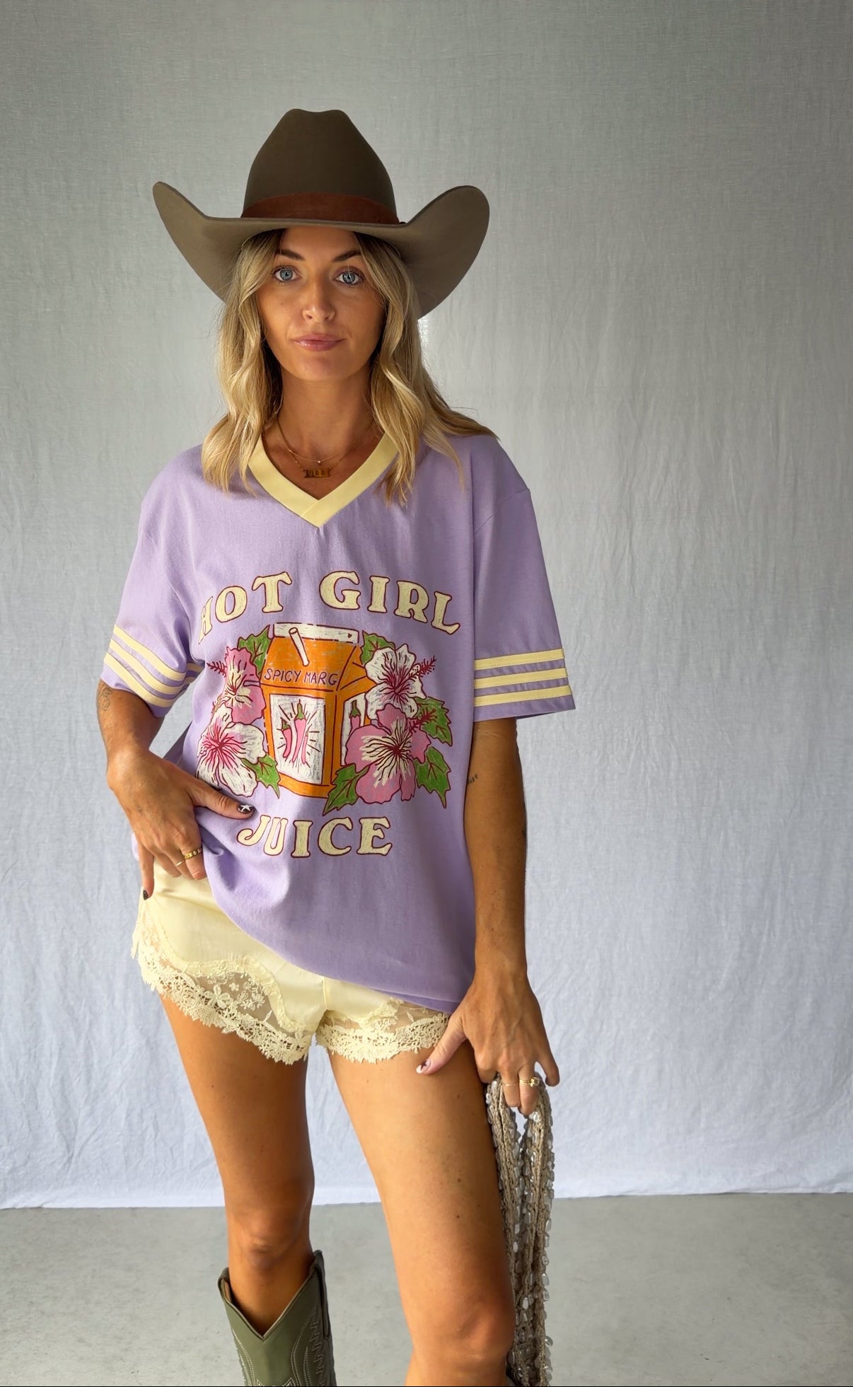 A woman modeling a lavender purple graphic t-shirt with yellow and white trim, paired with a brown cowboy hat, beige shorts, and green boots against a light gray backdrop.