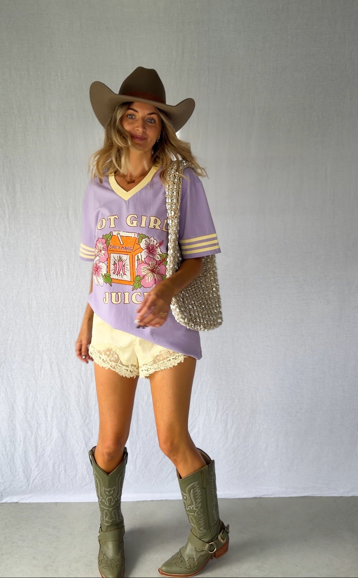 A woman modeling a lavender graphic t-shirt with 'Hot Girl Juice', paired with a brown cowboy hat, yellow lace shorts, olive green cowboy boots, and a silver pearl-beaded shoulder bag. Modern Coastal Cowgirl vibes.