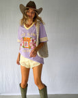 A woman modeling a lavender graphic t-shirt with 'Hot Girl Juice', paired with a brown cowboy hat, yellow lace shorts, olive green cowboy boots, and a silver pearl-beaded shoulder bag. Modern Coastal Cowgirl vibes.
