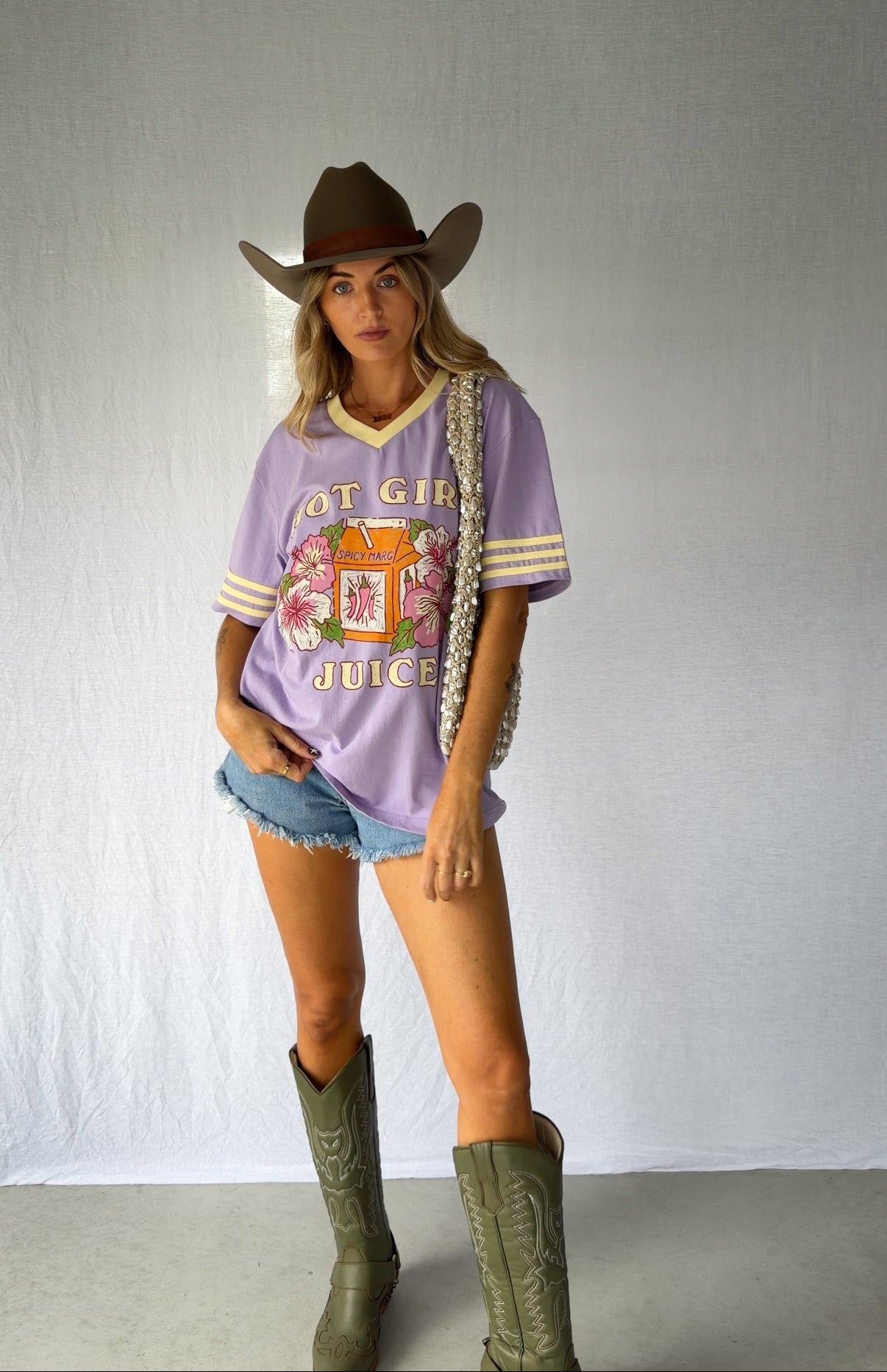 A woman modeling a lavender purple graphic t-shirt with a western theme, paired with denim shorts, a brown cowboy hat, and green cowboy boots. Modern coastal cowgirl. 