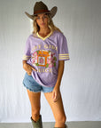 A woman modeling a lavender purple graphic t-shirt with yellow striped sleeves giving a modern jersey feel. Paired with denim shorts, a brown cowboy hat, and green cowboy boots. The perfect fun and bright tee for girls who love a spicy marg.