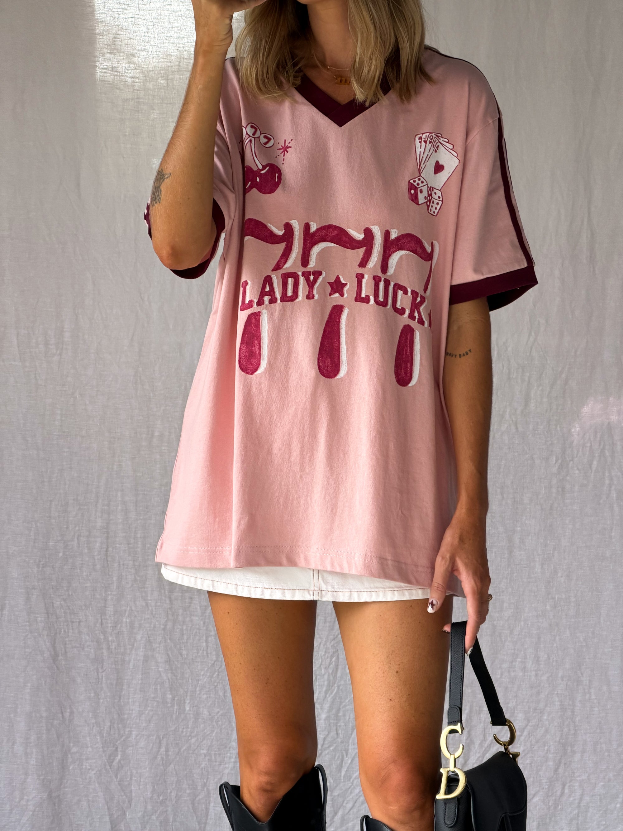 the Lucky Girl Tee is all about confidence, intuition, and calling in good things. Inspired by lucky motifs and the angel number 777, this women’s graphic tee blends playful symbolism with a bold, feminine edge. Pink base with burgundy trim for a flattering contrast. Jersey style vintage cotton.
The artist hand drawn art  western-style numbers and star detailing, finished with cherries, playing cards and dice graphics for a nostalgic, Americana-inspired feel.