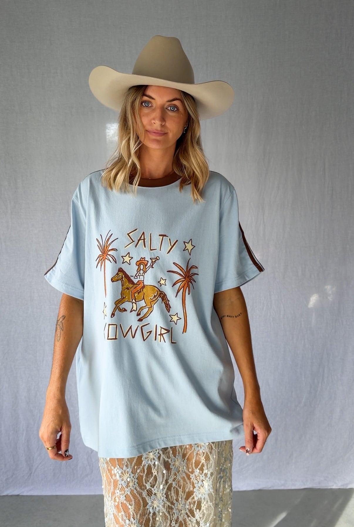 A woman wearing a light blue graphic t-shirt with a cowboy hat and a white lace and sequin skirt. The oversized tee features a hand-drawn graphic of a cowgirl in a shell bikini top riding a horse, palm trees, and the text 'SALTY COWGIRL'. For the modern salty cowgirl, this iconic tee is perfect for festival styling, rodeos and everyday wear all year round. 