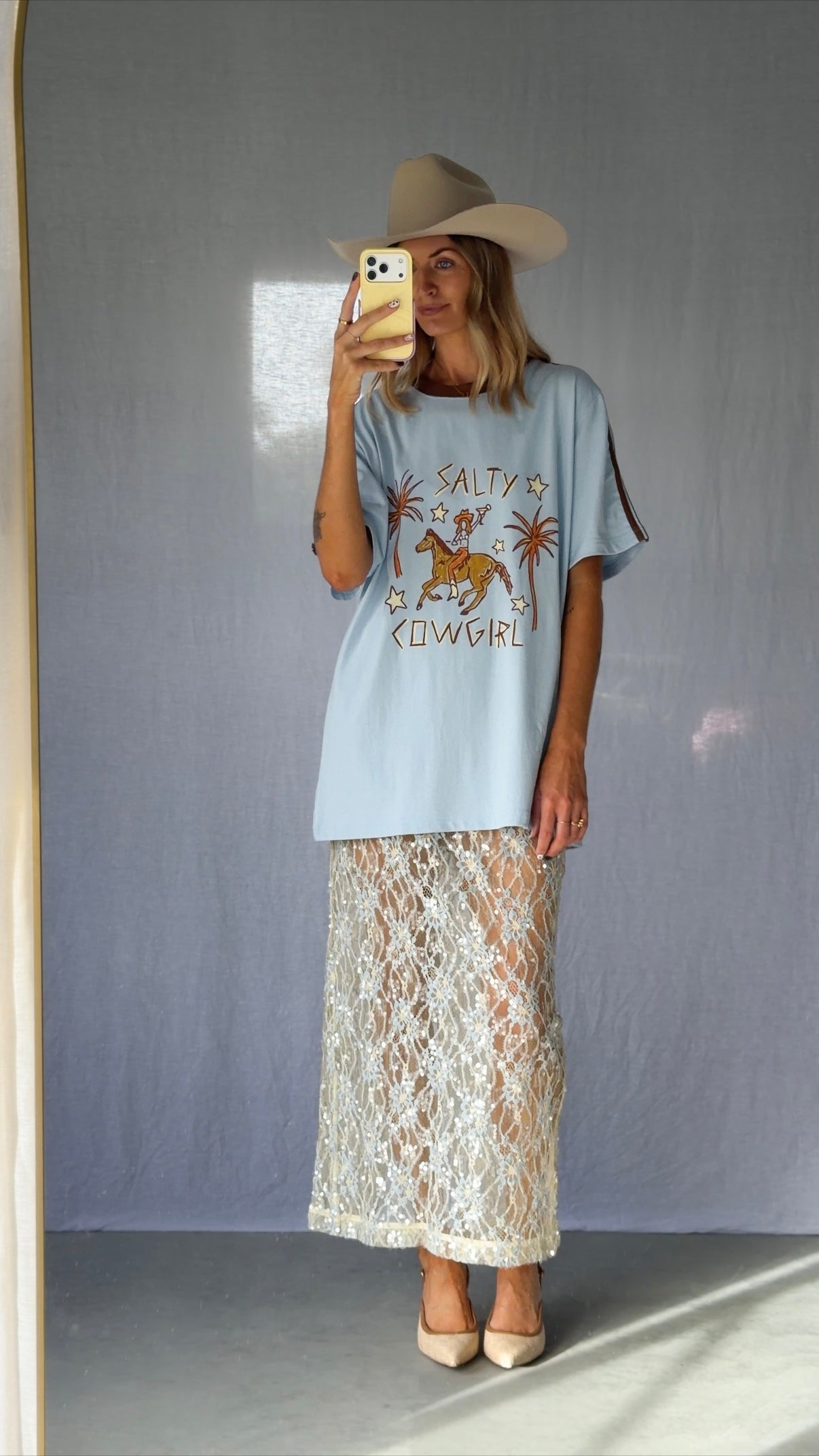 A woman wearing a light blue graphic t-shirt with a cowboy hat and a white lace and sequin skirt. The oversized tee features a hand-drawn graphic of a cowgirl in a shell bikini top riding a horse, palm trees, and the text 'SALTY COWGIRL'. For the modern salty cowgirl, this iconic tee is perfect for festival styling, rodeos and everyday wear all year round. 