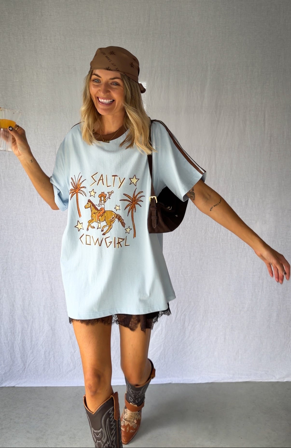 A woman wearing a light blue graphic t-shirt with a cowboy hat and a white lace and sequin skirt. The oversized tee features a hand-drawn graphic of a cowgirl in a shell bikini top riding a horse, palm trees, and the text 'SALTY COWGIRL'. For the modern salty cowgirl, this iconic tee is perfect for festival styling, rodeos and everyday wear all year round. 