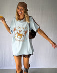 A woman wearing a light blue graphic t-shirt with a cowboy hat and a white lace and sequin skirt. The oversized tee features a hand-drawn graphic of a cowgirl in a shell bikini top riding a horse, palm trees, and the text 'SALTY COWGIRL'. For the modern salty cowgirl, this iconic tee is perfect for festival styling, rodeos and everyday wear all year round. 