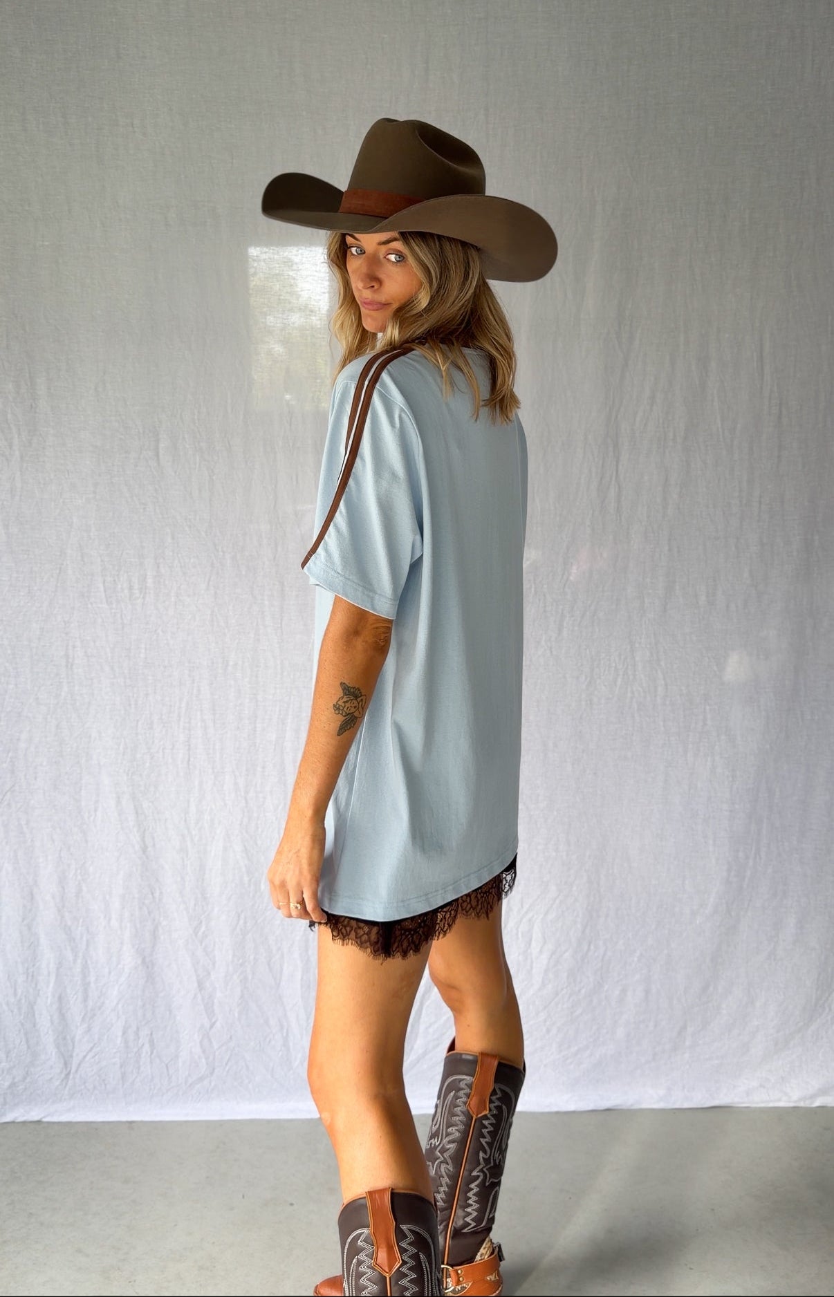 A woman wearing a light blue graphic t-shirt with a cowboy hat and a white lace and sequin skirt. The oversized tee features a hand-drawn graphic of a cowgirl in a shell bikini top riding a horse, palm trees, and the text 'SALTY COWGIRL'. For the modern salty cowgirl, this iconic tee is perfect for festival styling, rodeos and everyday wear all year round. 