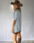 A woman wearing a light blue graphic t-shirt with a cowboy hat and a white lace and sequin skirt. The oversized tee features a hand-drawn graphic of a cowgirl in a shell bikini top riding a horse, palm trees, and the text 'SALTY COWGIRL'. For the modern salty cowgirl, this iconic tee is perfect for festival styling, rodeos and everyday wear all year round. 