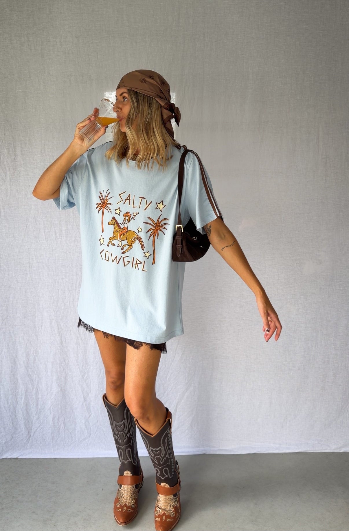 A woman wearing a light blue graphic t-shirt with a cowboy hat and a white lace and sequin skirt. The oversized tee features a hand-drawn graphic of a cowgirl in a shell bikini top riding a horse, palm trees, and the text 'SALTY COWGIRL'. For the modern salty cowgirl, this iconic tee is perfect for festival styling, rodeos and everyday wear all year round. 