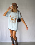 A woman wearing a light blue graphic t-shirt with a cowboy hat and a white lace and sequin skirt. The oversized tee features a hand-drawn graphic of a cowgirl in a shell bikini top riding a horse, palm trees, and the text 'SALTY COWGIRL'. For the modern salty cowgirl, this iconic tee is perfect for festival styling, rodeos and everyday wear all year round. 