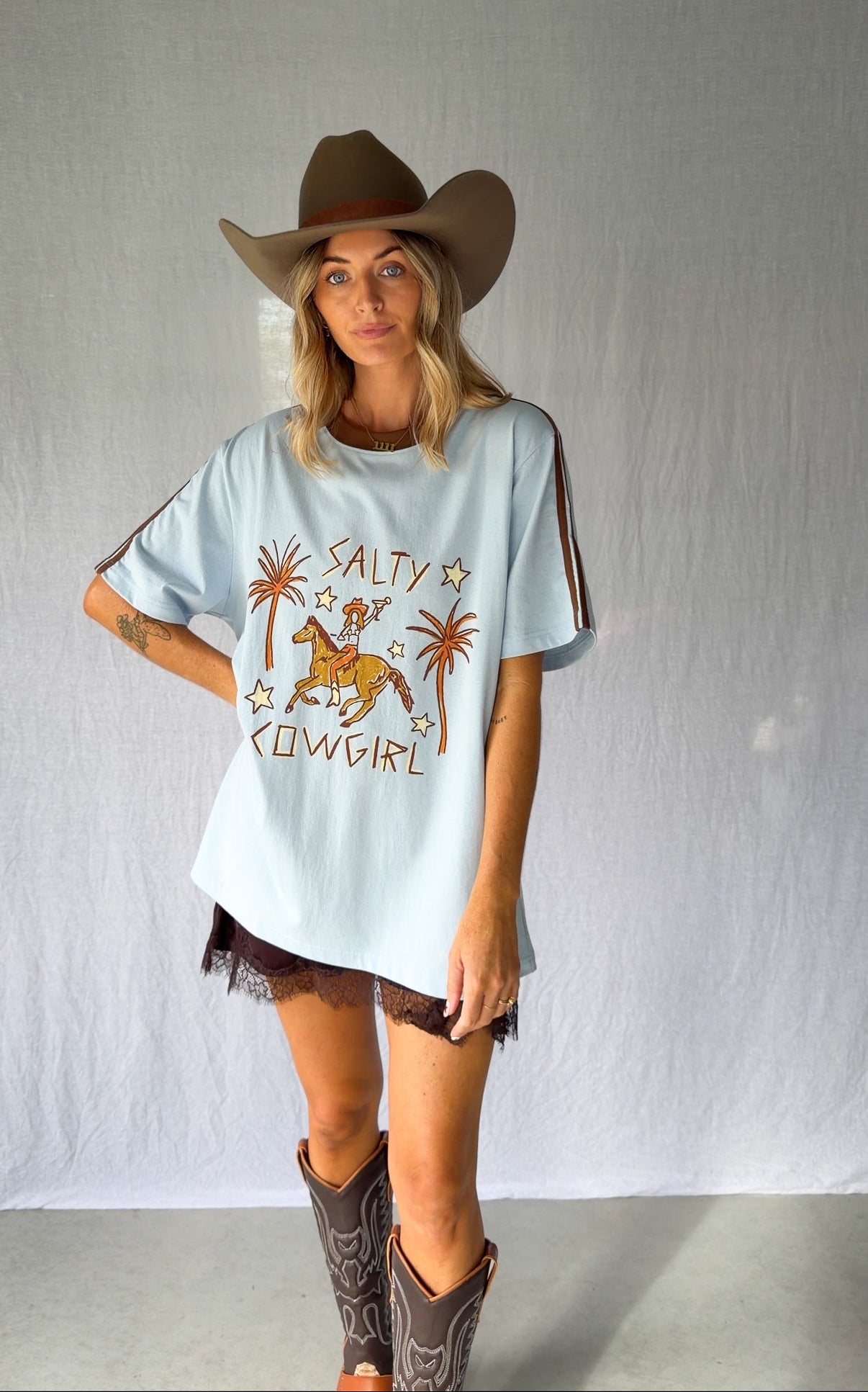 A woman wearing a light blue graphic t-shirt with a cowboy hat and a white lace and sequin skirt. The oversized tee features a hand-drawn graphic of a cowgirl in a shell bikini top riding a horse, palm trees, and the text 'SALTY COWGIRL'. For the modern salty cowgirl, this iconic tee is perfect for festival styling, rodeos and everyday wear all year round. 