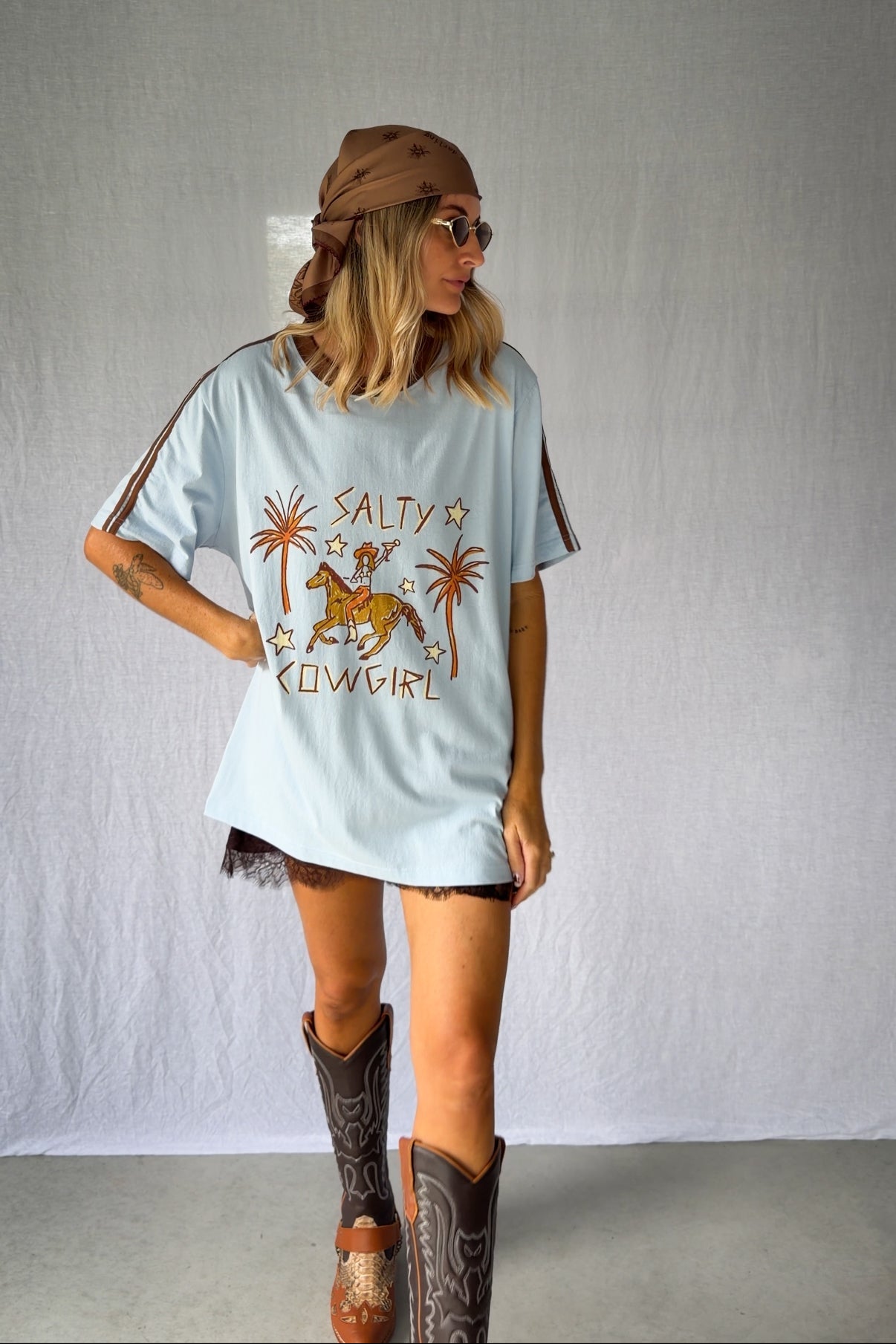 A woman wearing a light blue graphic t-shirt with a cowboy hat and a white lace and sequin skirt. The oversized tee features a hand-drawn graphic of a cowgirl in a shell bikini top riding a horse, palm trees, and the text 'SALTY COWGIRL'. For the modern salty cowgirl, this iconic tee is perfect for festival styling, rodeos and everyday wear all year round. 