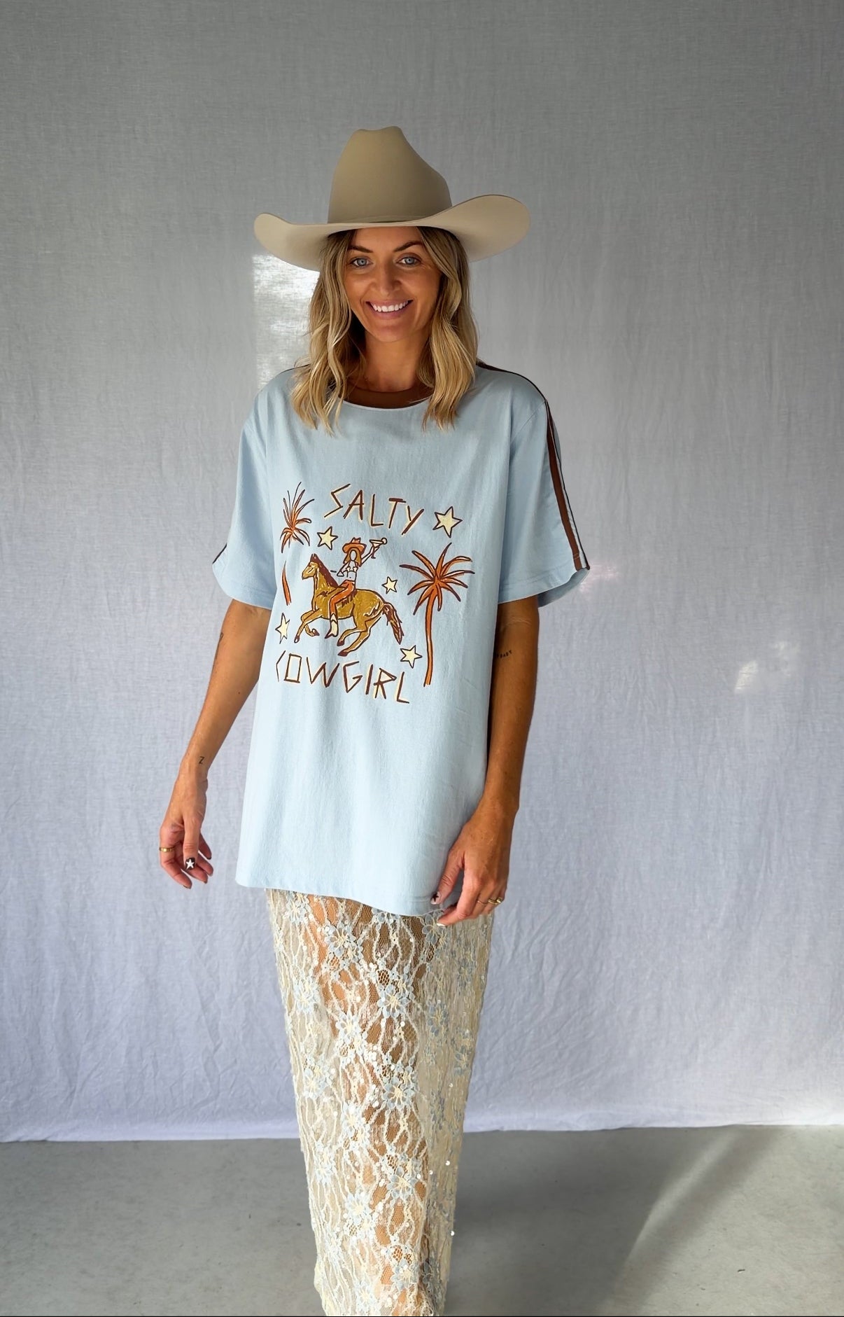 A woman wearing a light blue graphic t-shirt with a cowboy hat and a white lace and sequin skirt. The oversized tee features a hand-drawn graphic of a cowgirl in a shell bikini top riding a horse, palm trees, and the text 'SALTY COWGIRL'. For the modern salty cowgirl, this iconic tee is perfect for festival styling, rodeos and everyday wear all year round. 